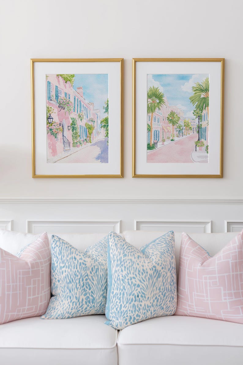 May include: Two framed watercolor prints of a street scene with pink buildings and blue shutters, hanging above a white couch with decorative pillows. The pillows are in shades of pink and blue.