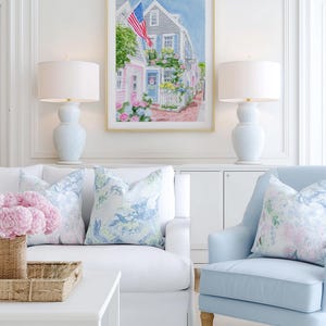 May include: A bright living room with a framed watercolour painting of a coastal town with an American flag. Two pale blue lamps sit either side of the artwork. A white sofa and a light blue armchair have floral cushions. A woven basket and white coffee table complete the scene.