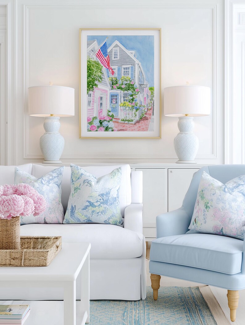 May include: A bright living room with a framed watercolour painting of a coastal town with an American flag. Two pale blue lamps sit either side of the artwork. A white sofa and a light blue armchair have floral cushions. A woven basket and white coffee table complete the scene.