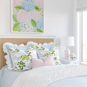 May include: A bed featuring a light blue and white gingham duvet, a white ruffled throw, and a cane headboard. The bed is styled with two large square pillows with a green and blue botanical print, a small pink diamond-patterned lumbar pillow, and a white ruffled sham.