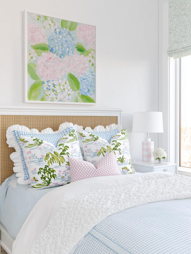May include: A bed featuring a light blue and white gingham duvet, a white ruffled throw, and a cane headboard. The bed is styled with two large square pillows with a green and blue botanical print, a small pink diamond-patterned lumbar pillow, and a white ruffled sham.