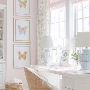 May include: Three framed butterfly art prints in pink, green, and blue, with gold frames, hang on a pink wall. A white desk with blue lamps sits near a window with pink blinds. A woven chair and basket add texture to the room.