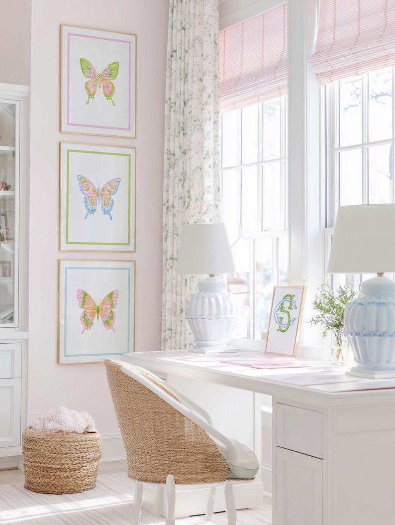 May include: Three framed butterfly art prints in pink, green, and blue, with gold frames, hang on a pink wall. A white desk with blue lamps sits near a window with pink blinds. A woven chair and basket add texture to the room.