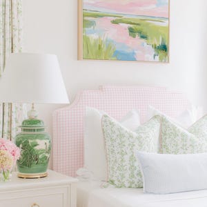 Op de afbeelding: Een slaapkamer met een roze gingham gestoffeerd hoofdeinde, wit beddengoed en sierkussens met groene bloemmotieven. Een groene keramische lamp met een witte kap staat op een wit nachtkastje naast een landschapsschilderij.
