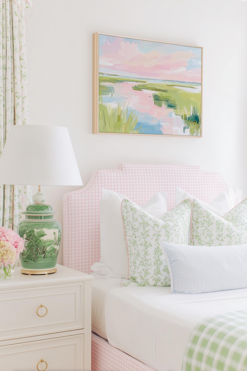 Op de afbeelding: Een slaapkamer met een roze gingham gestoffeerd hoofdeinde, wit beddengoed en sierkussens met groene bloemmotieven. Een groene keramische lamp met een witte kap staat op een wit nachtkastje naast een landschapsschilderij.