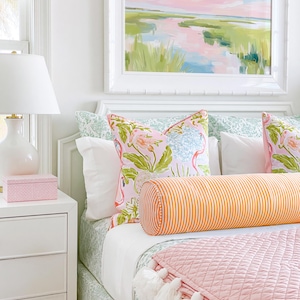 Op de afbeelding: Een slaapkamer met een bed voorzien van een groen bedsprei met patroon, twee kussens met flamingo-print, een lang oranje en wit gestreept rolkussen en een gewatteerde roze deken met witte kwastjes aan de onderkant.