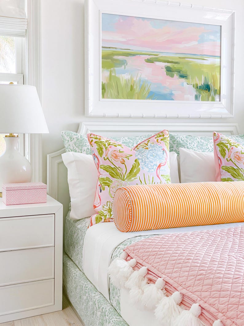 Op de afbeelding: Een slaapkamer met een bed voorzien van een groen bedsprei met patroon, twee kussens met flamingo-print, een lang oranje en wit gestreept rolkussen en een gewatteerde roze deken met witte kwastjes aan de onderkant.