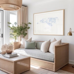 May include: A light grey sofa with a woven rattan frame and light grey cushions. The sofa is in a living room with a large framed print of the state of North Carolina with a blue floral pattern. The print is hanging on a white wall.