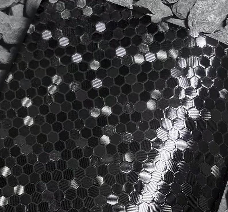 May include: A close-up of a black fabric with a repeating honeycomb pattern. The pattern is made up of small, six-sided shapes in varying shades of black and gray, creating a textured appearance. Some of the shapes have a reflective, shiny quality.