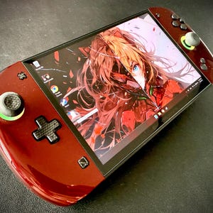 Könnte beinhalten: Handheld-Spielkonsole in glänzendem Burgunderrot. Der Bildschirm zeigt Anime-Kunst. Es hat ein schwarzes Steuerkreuz, Tasten und einen Joystick mit grünem Akzent. Das Design ist schlank und modern.