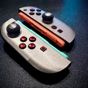 Könnte beinhalten: Zwei silberfarbene Gamecontroller mit roten und schwarzen Knöpfen. Ein Controller hat ein orangefarbenes Mittelteil und der andere ein hellblaues Mittelteil. Die Controller haben schwarze Joysticks.