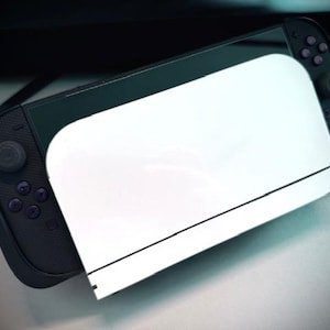 May include: A black handheld gaming console with purple buttons and a white screen protector. The console is partially visible, with the screen protector covering the screen. The background is blurred.