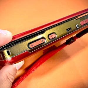 May include: A red and gold flip phone with a red wrist strap. The phone has a gold-colored frame with red buttons and a power button. The phone is held in a hand against an orange background.