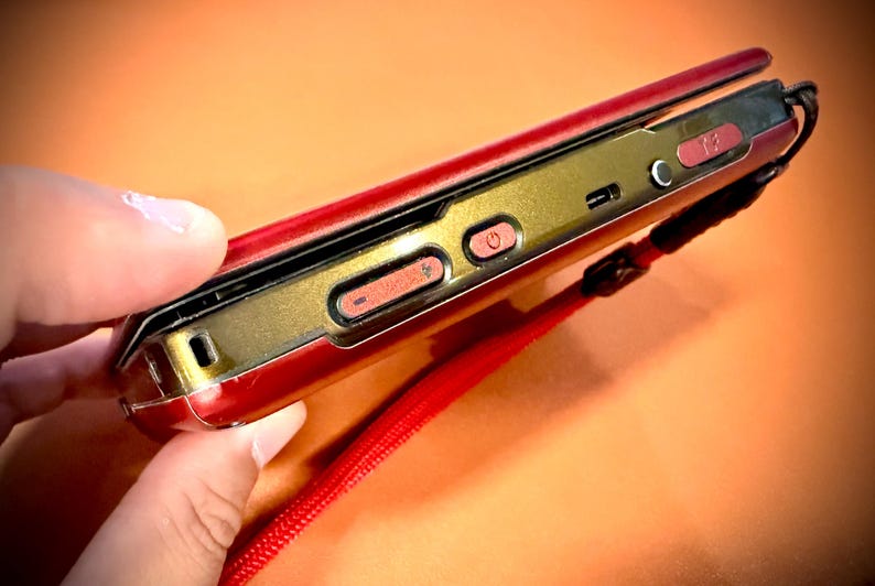 May include: A red and gold flip phone with a red wrist strap. The phone has a gold-colored frame with red buttons and a power button. The phone is held in a hand against an orange background.