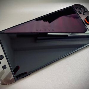 May include: Angled shot of a handheld gaming console with a large black screen. The console has gray sides with black buttons and a red joystick. The screen reflects a dark purple hue. The console is on a light gray surface.