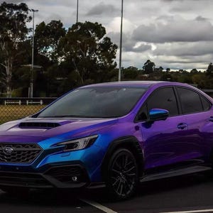May include: A Subaru WRX sedan with a color-shifting wrap that transitions from blue to purple. The car has black wheels and a black front grille. The background includes a cloudy sky and a grassy area.