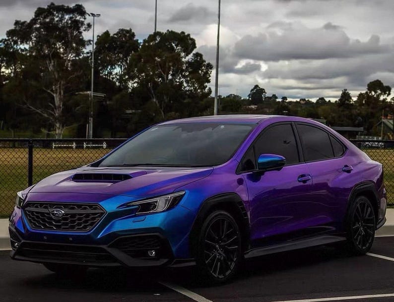 May include: A Subaru WRX sedan with a color-shifting wrap that transitions from blue to purple. The car has black wheels and a black front grille. The background includes a cloudy sky and a grassy area.