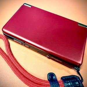 May include: A red handheld gaming console with a matching red wrist strap. The console is closed, showing a smooth, glossy surface. The wrist strap is red and braided, with a black and blue attachment.