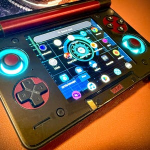 May include: A black handheld gaming device with a screen displaying app icons. It has red and black control buttons, including a directional pad and action buttons. The device has blue illuminated accents around the controls.
