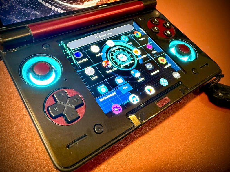 May include: A black handheld gaming device with a screen displaying app icons. It has red and black control buttons, including a directional pad and action buttons. The device has blue illuminated accents around the controls.