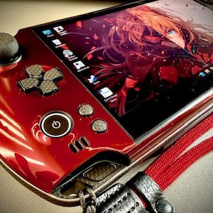 Puede incluir: Una consola de juegos portátil roja con una pantalla negra que muestra arte de anime. La consola tiene un joystick, un panel direccional y botones. Se adjunta un cordón rojo. La consola está sobre una superficie clara.