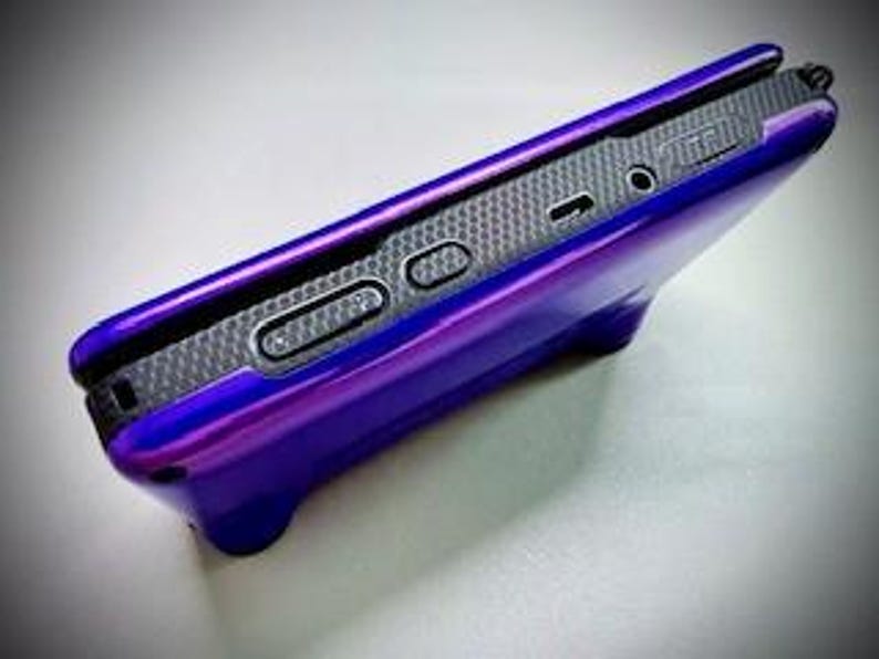 May include: A glossy purple phone case with a built-in stand. The case has a textured, dark gray interior and features buttons and ports along the side. The stand is integrated into the bottom of the case.