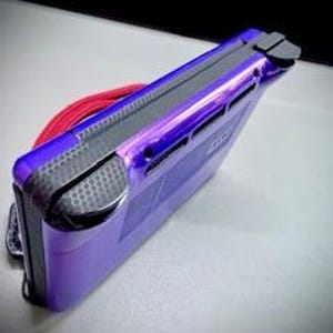 May include: A purple handheld gaming console with a black accent on the top and sides. A red cord is looped around the console. The console is on a white surface.