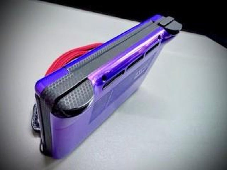 May include: A purple handheld gaming console with a black accent on the top and sides. A red cord is looped around the console. The console is on a white surface.