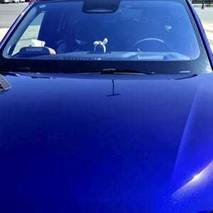 May include: A close-up shot of a shiny, deep blue car hood. The windshield reflects the surroundings, including the sky and buildings. The car's black side mirrors are visible, and a small decorative item is on the dashboard.