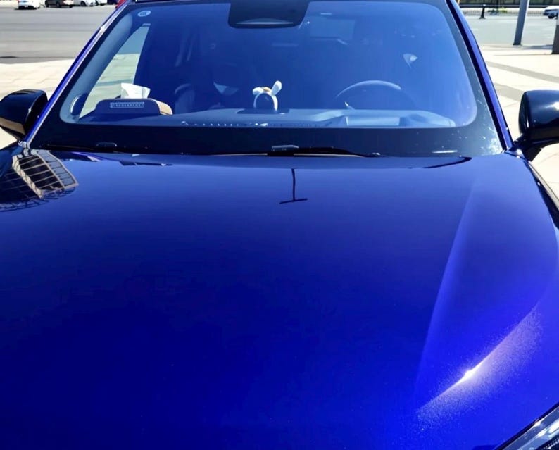 May include: A close-up shot of a shiny, deep blue car hood. The windshield reflects the surroundings, including the sky and buildings. The car's black side mirrors are visible, and a small decorative item is on the dashboard.