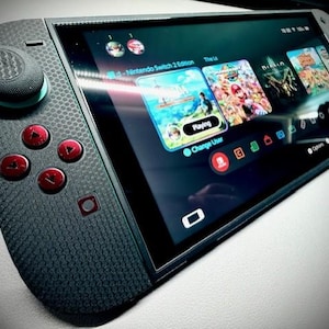 Könnte beinhalten: Handheld-Spielkonsole mit strukturierter grauer Oberfläche und roten Knöpfen. Der Bildschirm zeigt Spielsymbole, darunter "Nintendo Switch 2 Edition" und "Diablo". Die Konsole befindet sich auf einer weißen Oberfläche.