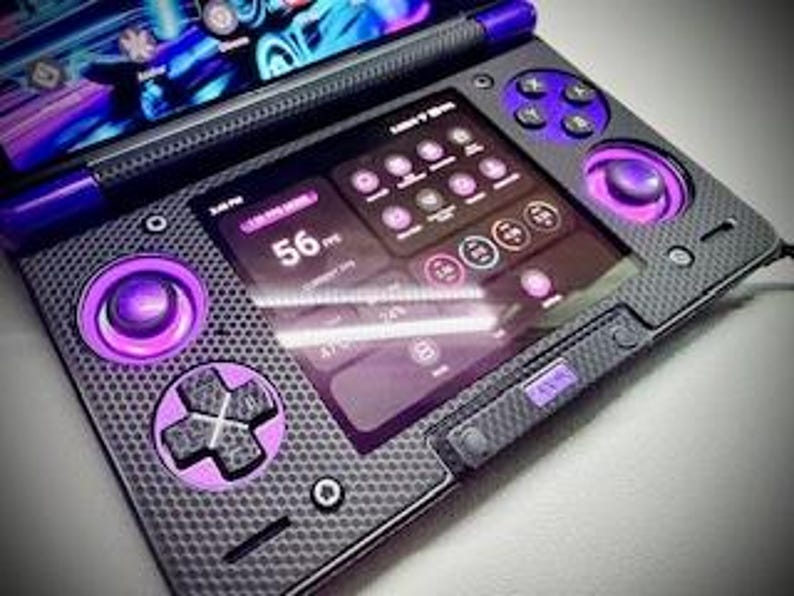 May include: A handheld gaming device with a dark gray carbon fiber texture and purple accents. The device features a screen displaying a user interface with various icons and data. It has purple control buttons and joysticks, and a stylus.