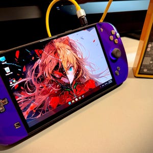 Può includere: Una console di gioco portatile viola con uno schermo che mostra opere d'arte anime. Il dispositivo ha un D-pad, pulsanti e levette analogiche. È collegato un cavo di ricarica giallo. Accanto c'è un dispositivo arancione con uno schermo che mostra il livello della batteria.