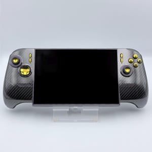 May include: A handheld gaming console featuring a dark gray carbon fiber patterned shell with a large black screen. The device includes gold-colored analog sticks, a gold directional pad, and four gold action buttons on the right side.