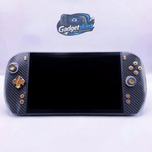 May include: A handheld gaming console featuring a black carbon fiber texture skin, gold-colored buttons, and a central black screen. Above the device, a logo displays the text Gadgetskinz with the tagline WRAP TECH IN STYLE in blue and white.