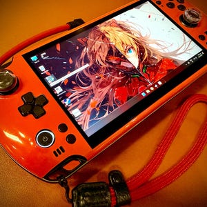 May include: Orange handheld gaming console with a screen displaying anime art. The console features a D-pad, buttons, and analog sticks. A red lanyard is attached. The screen shows a colorful anime character.