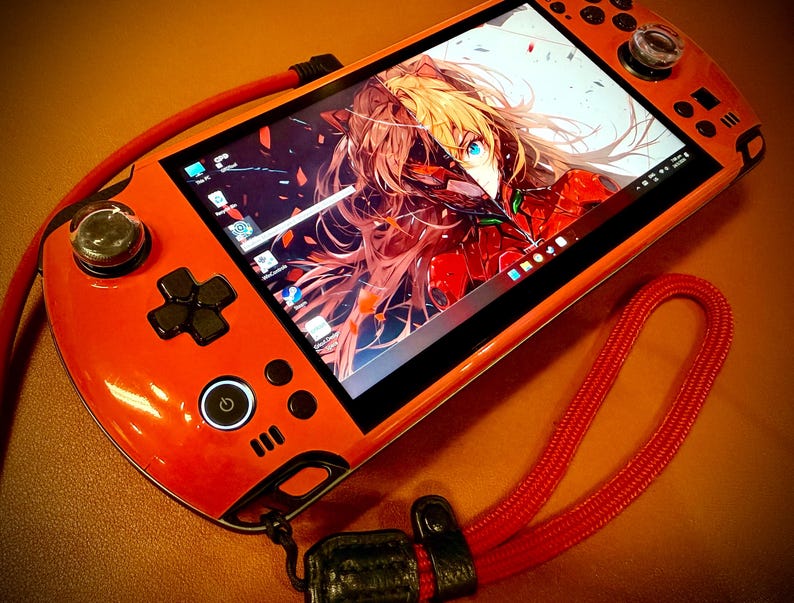 May include: Orange handheld gaming console with a screen displaying anime art. The console features a D-pad, buttons, and analog sticks. A red lanyard is attached. The screen shows a colorful anime character.