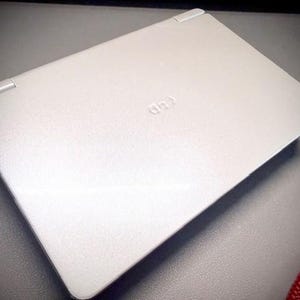 Puede incluir: Un portátil plateado con la tapa cerrada, con un logotipo sutil en relieve. El dispositivo tiene un diseño elegante y minimalista con esquinas redondeadas y una superficie texturizada. El portátil está sobre una superficie gris.