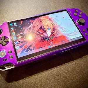 May include: A vibrant purple handheld games console, showing anime-style artwork on its screen. It has a D-pad, analogue sticks, and buttons. The device is on a dark surface.