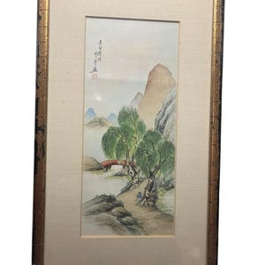 May include: Framed watercolour painting of a landscape with mountains, trees, and a small bridge. The artwork is in a gold-trimmed frame. The painting features soft colours and a serene scene, with what appears to be Asian characters in the upper left corner.