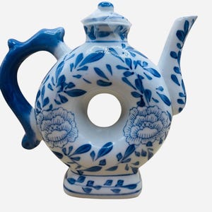 May include: A white ceramic teapot with a blue and white floral design. The teapot has a donut-shaped body with a hole in the center.