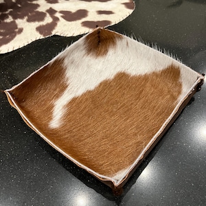 Op de afbeelding: Een vierkant dienblad van bruin leer met een witte koeienhuid voering. Het dienblad heeft een rustieke, westerse stijl.