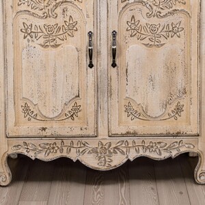 May include: A white painted wooden cabinet with carved floral details and distressed finish. The cabinet has two doors with ornate metal handles and a decorative base.