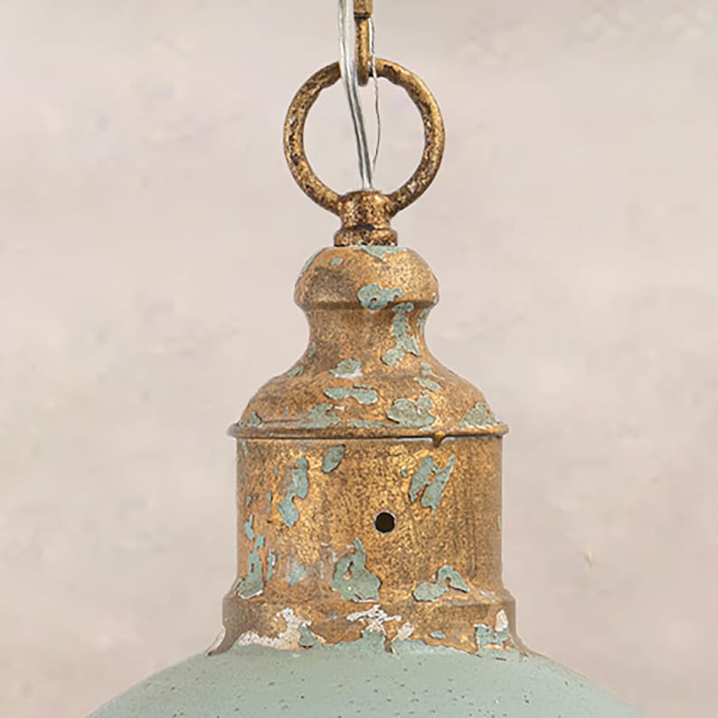 May include: Close-up of a vintage-style pendant light fixture. The fixture features a distressed gold finish with teal accents. A circular ring and chain are attached to the top for hanging. The light has a bell-shaped shade.