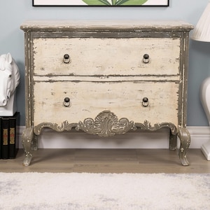 May include: A two-drawer chest with a distressed white and grey paint finish. The chest has ornate carvings on the front and black metal drawer pulls. The chest is sitting on a light wood floor.