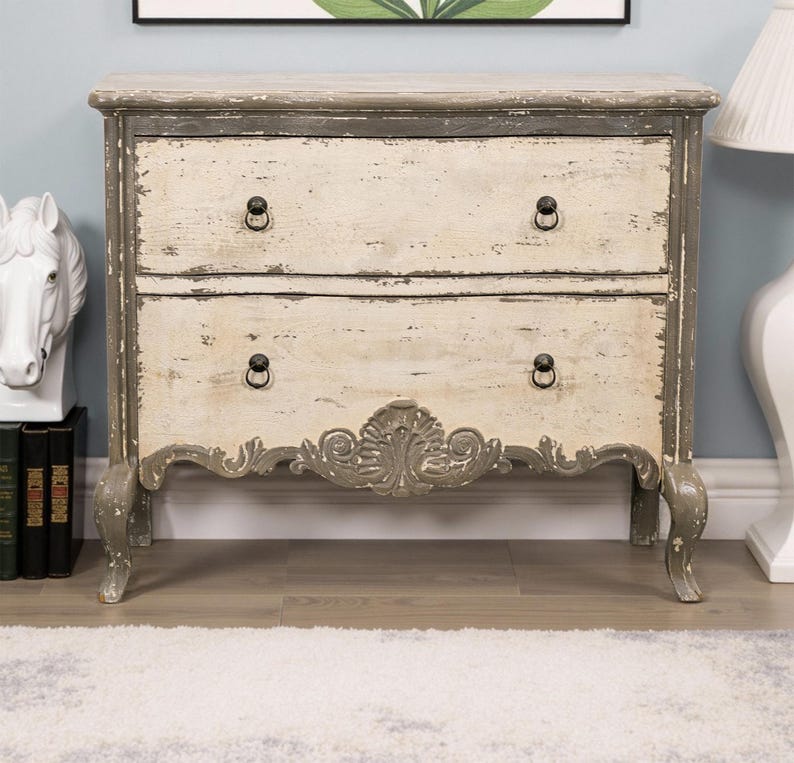 May include: A two-drawer chest with a distressed white and grey paint finish. The chest has ornate carvings on the front and black metal drawer pulls. The chest is sitting on a light wood floor.