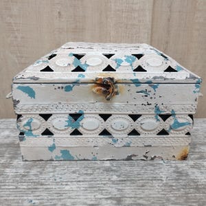 Puede incluir: Una caja de metal blanca con un diseño estampado en azul y blanco. La caja tiene una tapa con bisagras, un cierre de metal y un acabado desgastado.