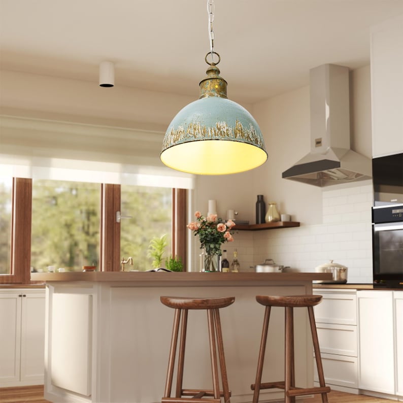 May include: A pendant light fixture with a distressed blue and gold finish hangs from a chain in a kitchen. The light has a rounded shade and illuminates the space below. The kitchen features a white island, wooden stools, and a window.
