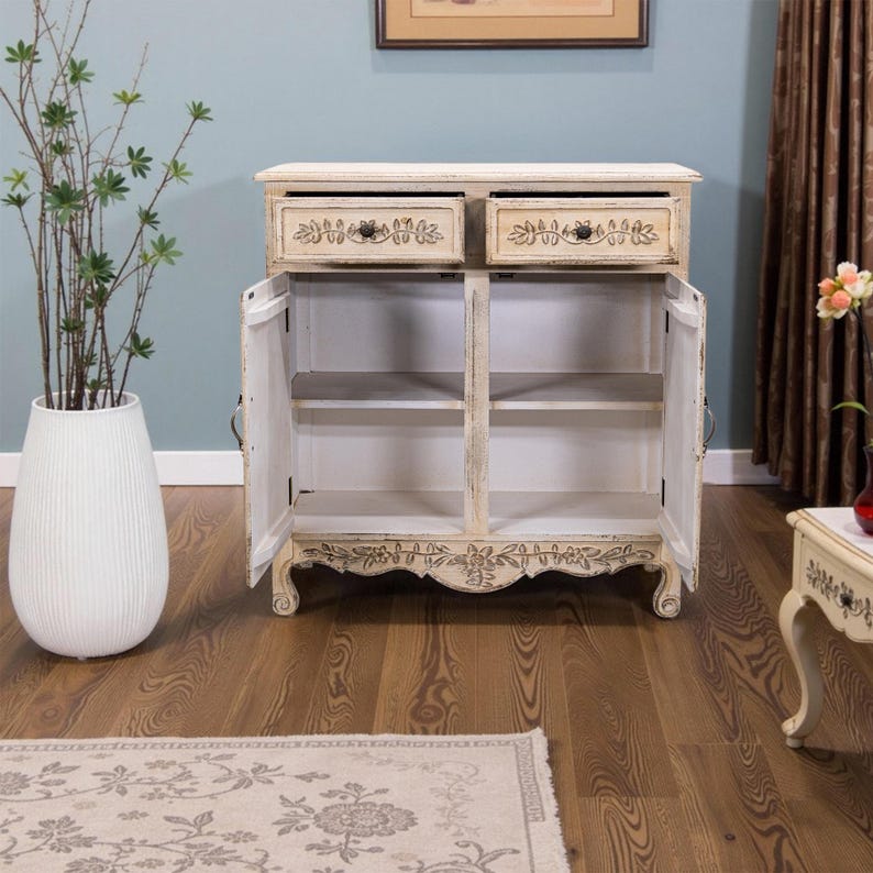 May include: White wooden cabinet with two drawers and two doors. The doors are open, revealing shelves inside. The cabinet has carved details and a distressed finish.