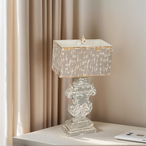 Puede incluir: Una lámpara de mesa gris y blanca envejecida con una pantalla rectangular. La base de la lámpara tiene un diseño tallado y ornamentado. La pantalla tiene un aspecto texturizado y desgastado con ribete dorado. La lámpara está sobre una superficie blanca.
